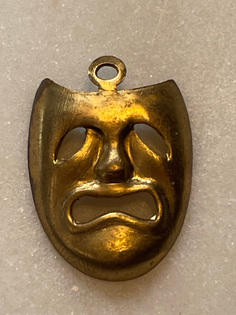 May include: A gold-colored metal pendant in the shape of a tragic mask. The mask has a sad expression with downturned eyes and a wide, open mouth.