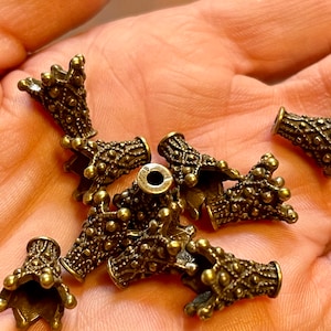 May include: A group of 10 antique gold-toned metal crown-shaped bead caps. The caps have a detailed design with a textured surface and a small hole at the top.