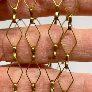 May include: Close-up of a gold-colored chain with a diamond-shaped pattern. The chain links are connected by small, round beads. The chain is held by a person's fingers, with a white background.