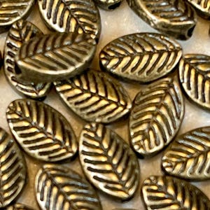 May include: A collection of gold-toned metal leaf-shaped beads. The beads have a textured surface with a raised vein pattern.