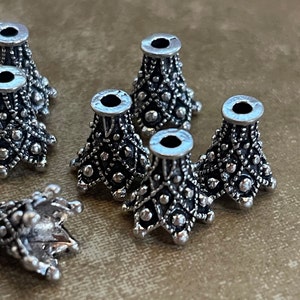 May include: Close-up of several silver-colored metal cone-shaped beads. Each bead has a detailed pattern of small dots and diamond shapes. The beads have a wide top opening and a decorative base.