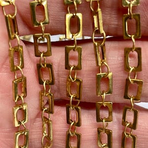 May include: Close-up of several strands of gold-colored chain. Each chain link is a rectangular shape with rounded corners. The chains are connected by small, round links. The background is blurred, with a hint of a hand holding the chains.