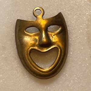 May include: A gold-colored metal pendant in the shape of a smiling theatrical mask. The mask has a large, open mouth and two small, round eyes.