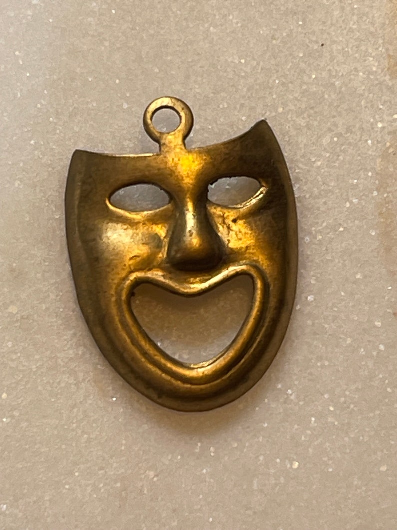 May include: A gold-colored metal pendant in the shape of a smiling theatrical mask. The mask has a large, open mouth and two small, round eyes.