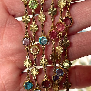 May include: A gold-tone chain necklace featuring alternating circular links with faceted multi-colored glass stones and textured gold-tone flower-shaped metal charms. The stones appear in shades of light green, blue, pink, and deep purple.