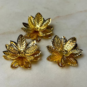 May include: Three gold-colored metal flower-shaped components. Each component has multiple detailed petals and a central hole. The components are arranged on a light-colored, marble-like surface.