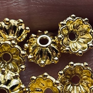 May include: Close-up of six gold-toned metal bead caps with a decorative, scalloped design. The bead caps have a small hole in the center for threading.