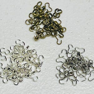 May include: A pile of silver and gold jump rings. The jump rings are in the shape of a heart with a hook on one end. The jump rings are used for jewelry making.