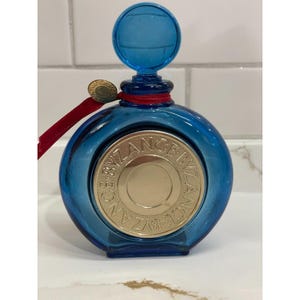 May include: A round, blue glass perfume bottle with a matching stopper. The bottle features a gold-colored medallion with the text "CE BYZANCE" around the edge. A red ribbon and a small gold coin are attached to the bottle.