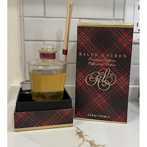 May include: A Ralph Lauren fragrance diffuser with a clear glass bottle containing a yellow liquid. The bottle has a red stopper and is accompanied by wooden reeds. The box is red and black plaid with gold trim and the text "RALPH LAUREN Fragrance Diffuser".
