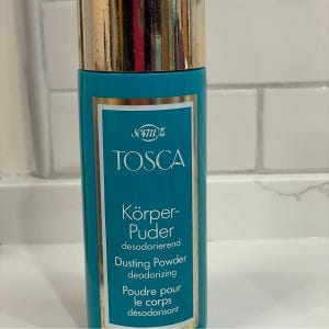 May include: A teal dusting powder bottle with a gold cap. The label reads "TOSCA" and "Dusting Powder deodorizing". The bottle is cylindrical and appears to be made of glass or a similar material. The background is a white tiled wall.