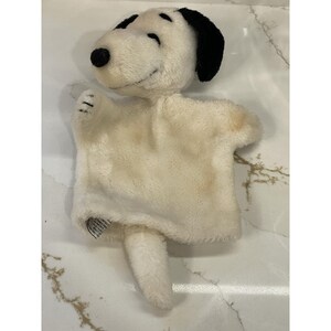May include: A Snoopy hand puppet made of soft, off-white plush material. The puppet features Snoopy's iconic black ears, nose, and eyebrows. The puppet has a small tail and is designed to fit over the hand for play.