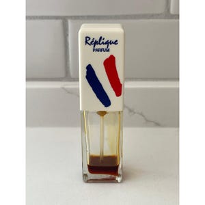 May include: A rectangular perfume bottle with a white cap featuring the text "Réplique PARFUM" in blue. The cap has blue and red diagonal stripes. The bottle contains a dark amber liquid.