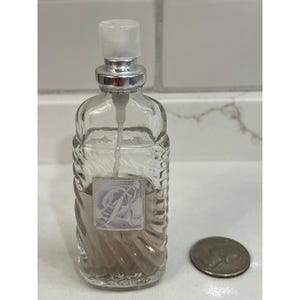 May include: A clear glass perfume bottle with a textured, ridged design. The bottle has a silver-colored sprayer and a label with a floral design and the letter "R". A quarter is next to the bottle.