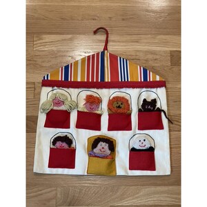 May include: A fabric wall hanging with a striped top and red trim. It features eight pockets, each holding a felt puppet with yarn hair or fur. The puppets include a girl, a boy, a lion, a cat, and others. The background is white.