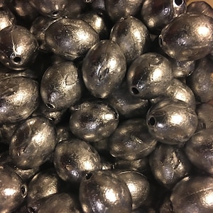 May include: A close-up shot of a pile of silver-colored fishing weights. The weights are oval-shaped with a hole at one end. The weights have a shiny, metallic finish and are densely packed together.