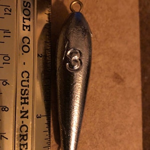 May include: A silver fishing sinker with a brass loop at the top and bottom. The sinker is shaped like a bullet and has the number "3" stamped on the side. The sinker is about 2 inches long.