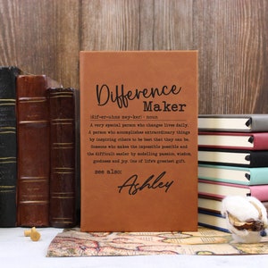 May include: A brown leather journal with the text "Difference Maker" and a definition of the word. The text "see also: Ashley" is printed below the definition.