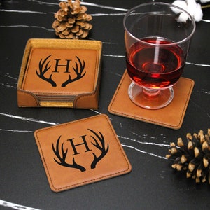 May include: Set of four brown leather coasters with a black deer antler design and the letter "H" in the center. The coasters are stacked on top of each other and are sitting on a black surface.