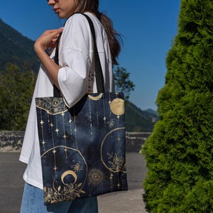 May include: A dark blue tote bag featuring a celestial design with gold moons, stars, and floral accents. The bag has black straps and is being carried outdoors. The background includes mountains and a blue sky.