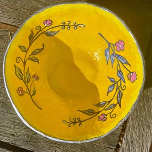 Może przedstawiać: Żółta ceramiczna miska z malowanym wzorem kwiatowym w kolorze czarnym, niebieskim i różowym. Miska jest ozdobiona wieńcem ze stylizowanych liści i kwiatów.