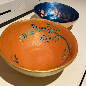 May include: A hand-painted orange bowl with a gold rim and a green and blue floral design. The bowl is decorated with gold dots.