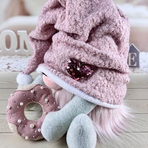 May include: A plush gnome doll with a pink textured hat adorned with a sequined heart. The doll has a pink beard, blue arms and legs, and is holding a pink donut decorated with pearls. The overall theme is whimsical and decorative.