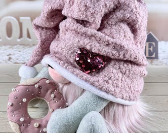 Può includere: Una bambola gnomo di peluche con un cappello rosa strutturato adornato da un cuore di paillettes. La bambola ha una barba rosa, braccia e gambe blu e tiene in mano una ciambella rosa decorata con perle. Il tema generale è stravagante e decorativo.