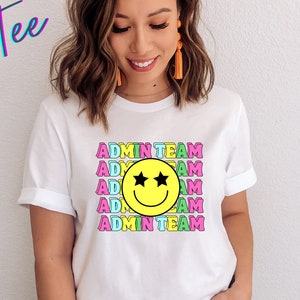 May include: White t-shirt with a colorful graphic design featuring a smiley face with stars for eyes and the text "ADMIN TEAM" repeated multiple times.