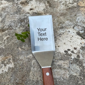 Puede incluir: Una espátula de acero inoxidable con mango de madera. La hoja de la espátula tiene el texto "Your Text Here" impreso. El mango es de color marrón oscuro y está asegurado con dos remaches plateados.