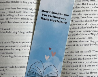 May include: A blue bookmark with a white open book illustration and the text "Don't Bother Me I'm Visiting My Book Boyfriend". The bookmark has a light blue background with white clouds and pink hearts.