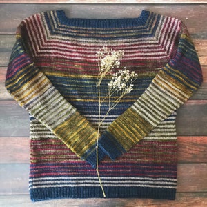 May include: A striped sweater in shades of blue, red, brown, and yellow. The sweater has a boat neck and long sleeves. A sprig of dried flowers is placed in front of the sweater.