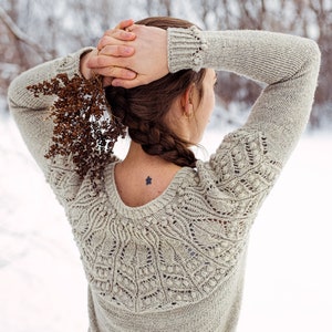 May include: A woman wearing a light brown knitted sweater with a delicate lace pattern. The sweater has a scoop neck and long sleeves. The woman is standing in a snowy landscape.
