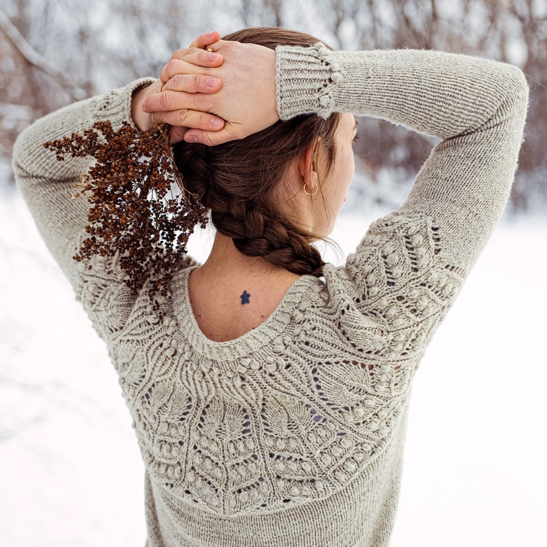 May include: A woman wearing a light brown knitted sweater with a delicate lace pattern. The sweater has a scoop neck and long sleeves. The woman is standing in a snowy landscape.