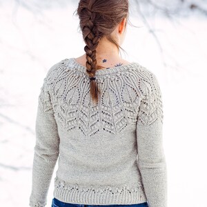 May include: A woman wearing a light gray knitted sweater with a delicate lace pattern and blue jeans. Her hair is braided and pulled back.