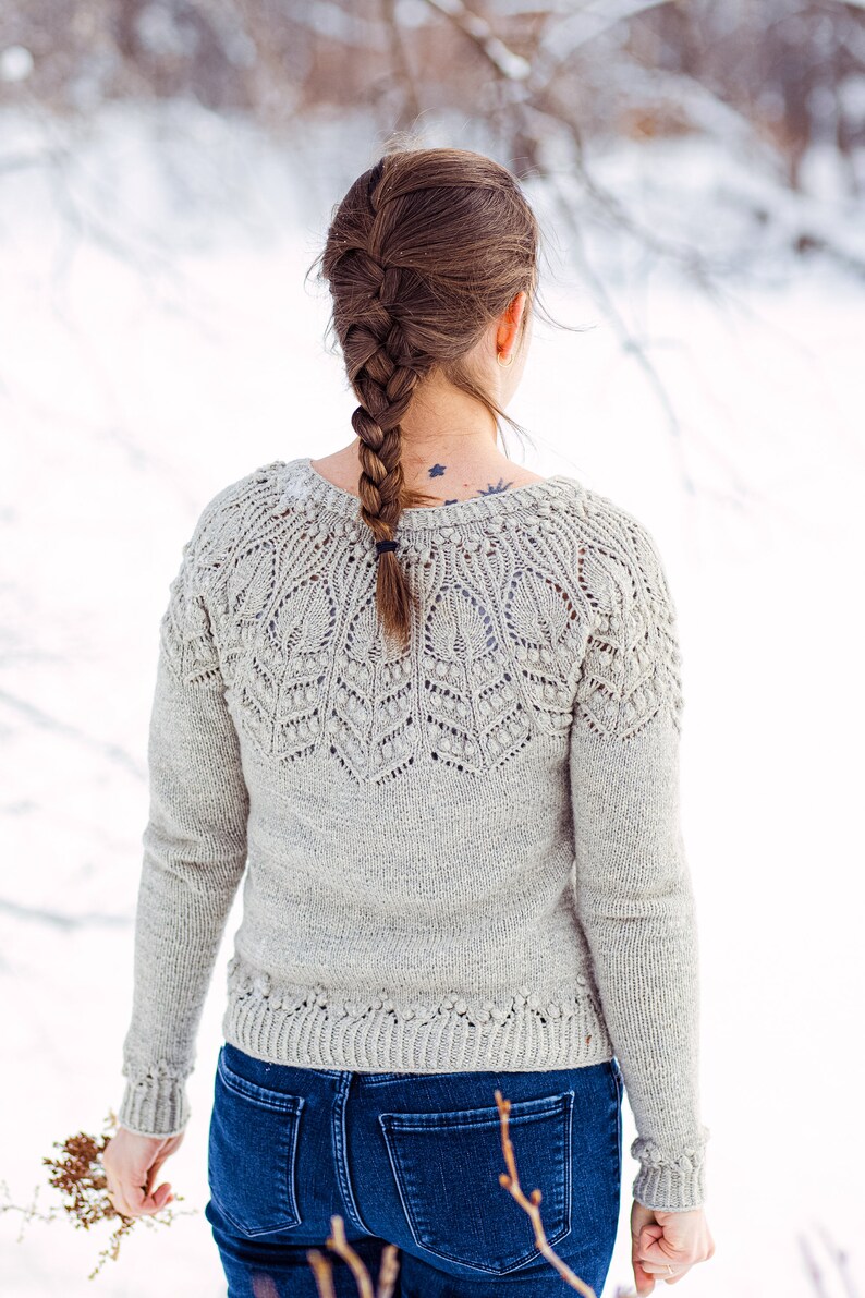 May include: A woman wearing a light gray knitted sweater with a delicate lace pattern and blue jeans. Her hair is braided and pulled back.