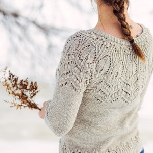 May include: A woman wearing a light beige knitted sweater with a delicate lace pattern. The sweater has a round neckline and long sleeves. The woman is standing in a snowy landscape.