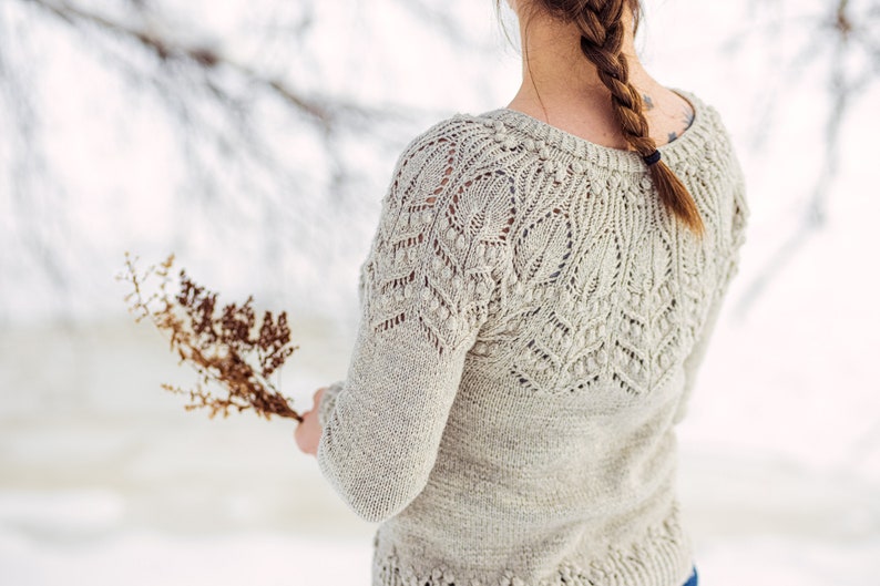 May include: A woman wearing a light beige knitted sweater with a delicate lace pattern. The sweater has a round neckline and long sleeves. The woman is standing in a snowy landscape.