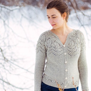 May include: A woman wearing a light gray knitted cardigan with a delicate lace pattern and button closure. The cardigan has a V-neck and long sleeves. The woman is standing in a snowy outdoor setting.
