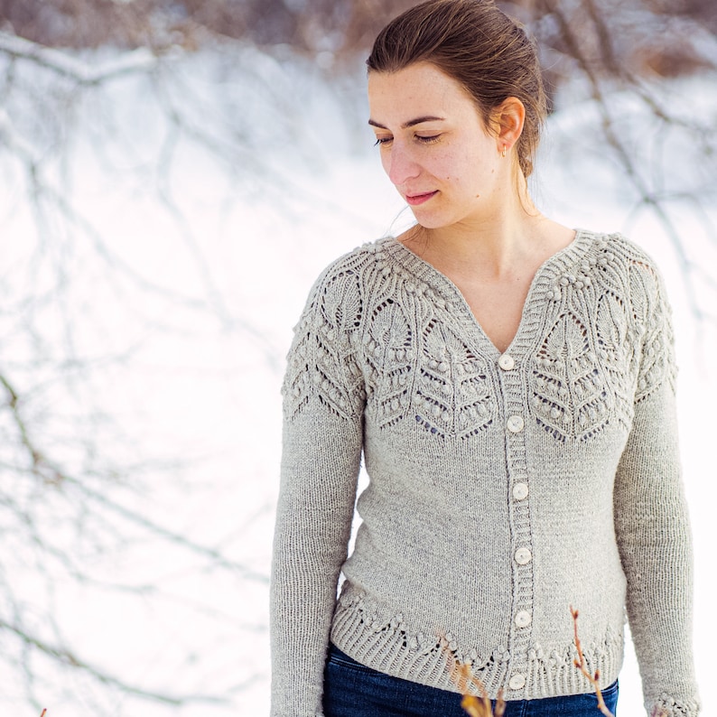 May include: A woman wearing a light gray knitted cardigan with a delicate lace pattern and button closure. The cardigan has a V-neck and long sleeves. The woman is standing in a snowy outdoor setting.