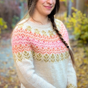 May include: A white knitted sweater with a colorful geometric pattern in shades of pink, orange, and green. The sweater has a crew neck and long sleeves.