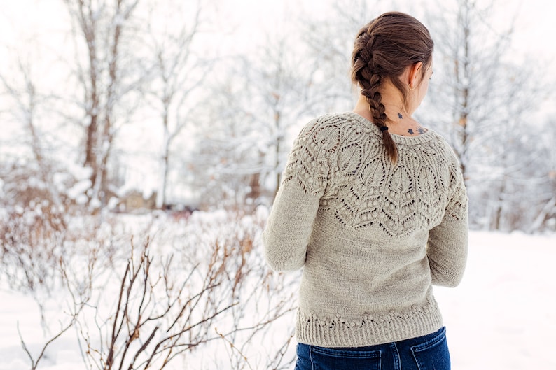 May include: A woman wearing a light gray knitted sweater with a delicate lace pattern. The sweater has a v-neck and long sleeves. The woman is standing in a snowy landscape.