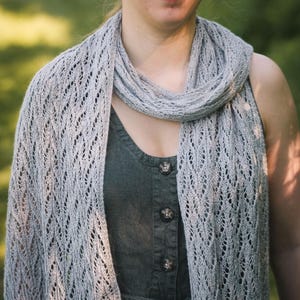 Può includere: Uno scialle lavorato a maglia grigio chiaro caratterizzato da un intricato motivo in pizzo traforato con colonne verticali di occhielli. Lo scialle è drappeggiato attorno al collo e alle spalle sopra un abito smanicato grigio antracite scuro.
