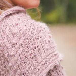Peut inclure: Un pull en tricot rose poudré présentant un motif texturé de torsades verticales, des détails en dentelle ajourée délicats et de petites boules en relief. Le vêtement a une texture douce et duveteuse avec une couture d'épaule structurée.