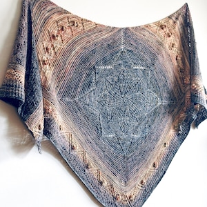 May include: A handcrafted, square shawl with a starburst design in the center. The shawl features a gradient of colors, transitioning from blue to tan and brown. Decorative beads are placed along the edges, adding texture and detail to the knitted accessory.