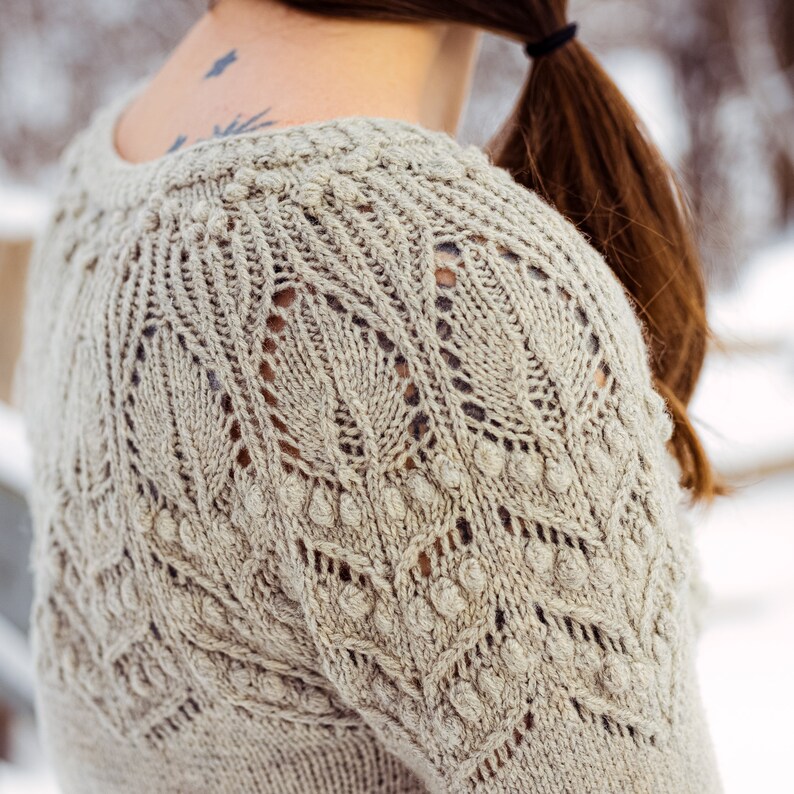 May include: A close-up of a beige knitted sweater with a delicate lace pattern and textured bobbles. The sweater has a round neckline and a relaxed fit.
