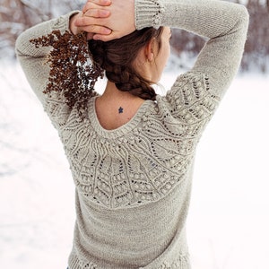 May include: A woman wearing a light gray knitted sweater with a delicate lace pattern on the back. The sweater has a round neckline and long sleeves. She is wearing blue jeans and has her hair in a braid.