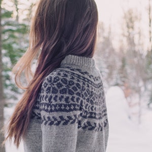 Può includere: Una donna con lunghi capelli castani indossa un maglione grigio e nero con motivo. Il maglione ha un design a intarsio.