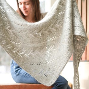 Peut inclure: Un châle tricoté à la main, de couleur gris clair, avec des motifs ajourés complexes. Le châle est de forme triangulaire et semble doux et léger. Le design délicat présente un motif géométrique répétitif, créant un aspect texturé et aéré. Le châle est tenu, montrant sa forme complète.
