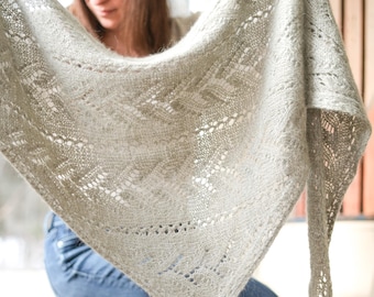 May include: A pale grey, hand-knitted shawl with detailed openwork patterns. The shawl is triangular and appears soft and lightweight. The delicate design features a repeating geometric motif, creating a textured and airy appearance. The shawl is held up, showcasing its full form.